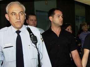 A man is being taken into a courthouse wearing a black shirt. He has brown hair. He is being escorted by an elderly man with white hair in a button-down.