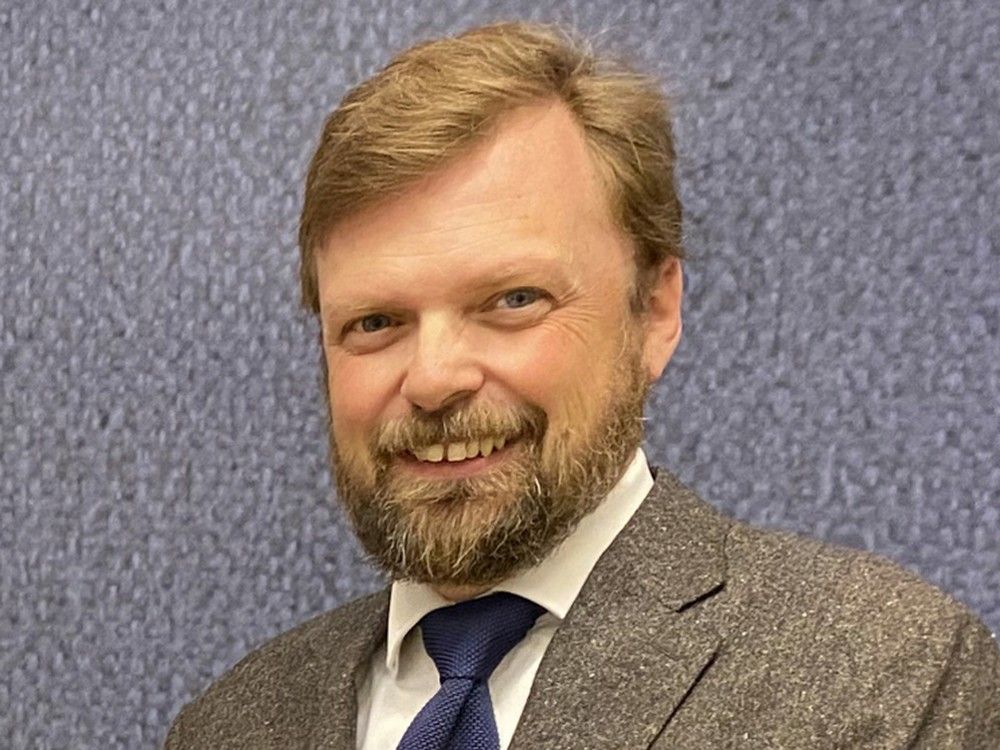 A bearded nab in a suit and tie smiles at the camera in front of a patterned background.