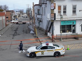 A police vehicle and crime-scene tape are at an intersection, with a white building in the frame. It has businesses on the first floor and residences on the second storey.