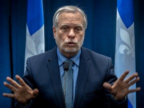 Quebec's public health director, dr luc boileau, is seen gesturing with both hands up in a blue suit in front of quebec flags at a press conference.