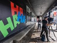 A TV reporter and camera operator stand outside a building with 'HA HA HA' painted on it