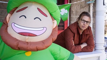 A woman leans on a porch rail, looking serious. In the foreground is a blowup St. Patrick's leprechaun.