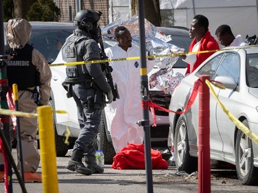 An officer in SWAT gear and a paramedic in a hazmat suit stand near three men by parked cars.