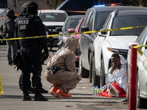 A paramedic kneels to speak with a man sitting against a parked car. A police officer looks on.