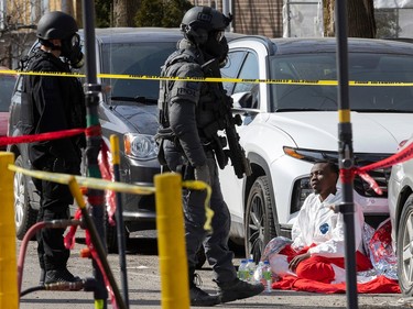 Police in SWAT gear stand near parked cars. A man is sitting on the ground in front of them.