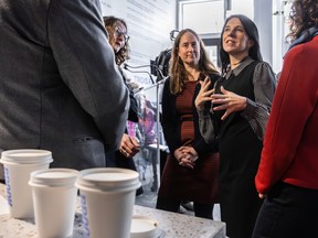 A woman with black hair (Montreal Mayor Valerie Plante) is seen in the background with three coffee cups on the table to the left in the foreground. There are other people to the right and left of the mayor but she is the focus.