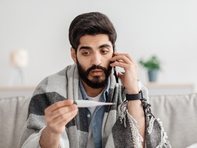 A man with a cellphone to his ear looks at a thermometer. He is wrapped in a blanket.