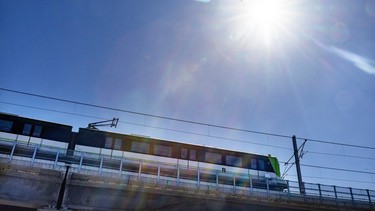 An elevated train against a blue sky. A bird is flying through rays of sun.