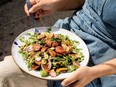 a colourful fattoush salad is seen sitting in a white plate, which is resting on a person's lap in close up. The person's hands are on either side of the plate, and a blue apron over their belly is visible.