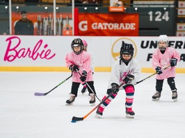 Three elementary-school-age girls are wearing pink and white gear while playing on a hockey rink.