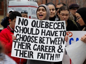 A woman wearing a hijab holds a sign that reads 'No Quebecer should have to choose between their career & their faith!!!'