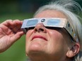 A closeup of a white-haired woman staring up at the sky, holding a pair of eclipse glasses that are covering her eyes.