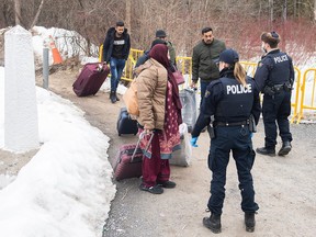 Police carrying suitcases next to snowbanks are stopped by police officers on a pathway.