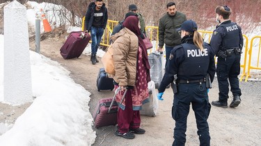 Police carrying suitcases next to snowbanks are stopped by police officers on a pathway.