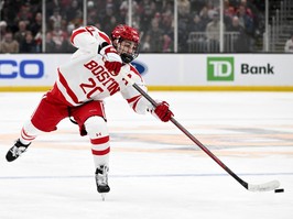 Boston University's Lane Hutson is seen in the team's white uniform shooting the puck.