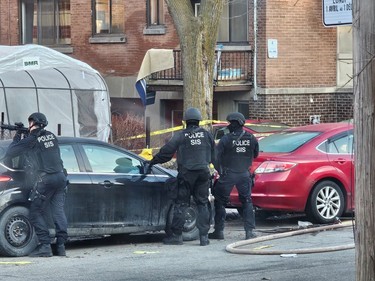 Three police officers next to cars parked on a residential street. One officer is leaning on a car with a large gun.