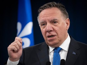 François Legault gestures while speaking at a microphone with a Quebec flag behind him