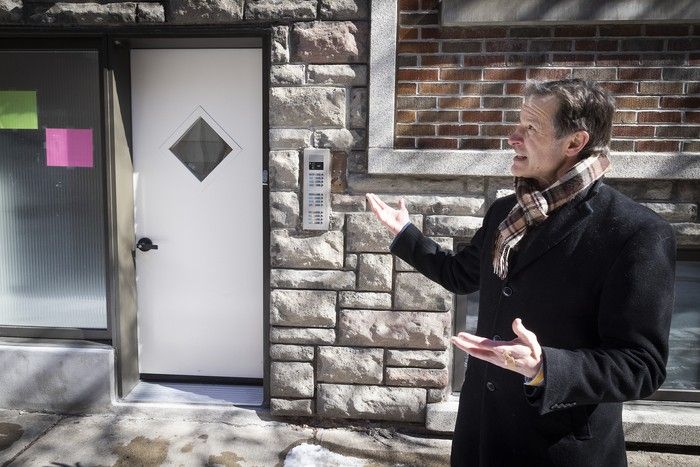 A man in a coat and scarf gestures toward the door of a building.