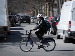 A man dressed in dark colours wearing a white helmet is riding a bixi bike-share across a street with cars visible on either side.