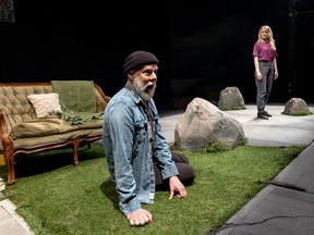 A man strikes a dog-like pose while kneeling on a stage made to look like grass. A couch is in the background, and a woman stands amid rocks on the other side of the stage.
