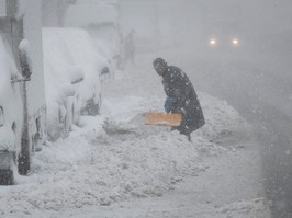 A person shovels snow while it's still falling. There are headlights in the distance behind them.