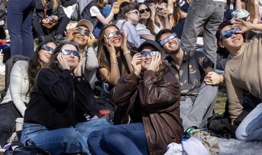 A group of people wear eclipse glasses while smiling and looking up.