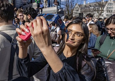 A woman holds eclipse glasses and her phone while staring up in a crowd of people.
