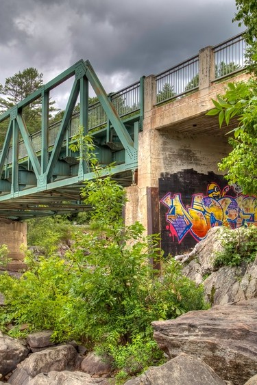A steel bridge seen from below, with bright graffiti on one of the concrete supports.