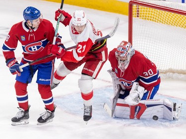 Canadiens' Lane Hutson has his stick in the armpit of Red Wings' Lucas Raymond in front of the Canadiens crease, while the puck is in the air in front of Cayden Primeau's pad