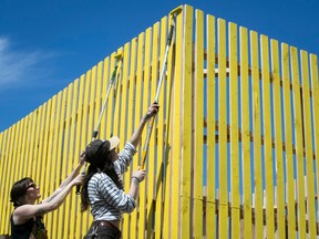 Two women paint a tall yellow fence.