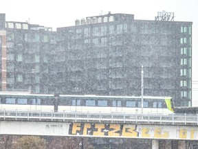 A REM train travels on a graffiti-tagged elevation amid snowfall, with buildings in the background.