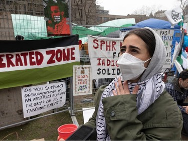 A young woman looks up holding her hand on her kaffiyeh next to a fence with signs on it at an encampment downtown