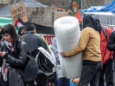 People gather outside a fence with banners attached to it, one of them holding a large rolled up piece of foam