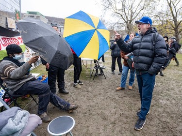 A man in a blue baseball cap points a finger to gesture as he speaks to others, some holding umbrellas open toward him
