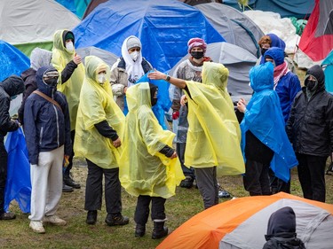 About a dozen people gather in a circle wearing plastic rain ponchos as one gestures amid camping tents