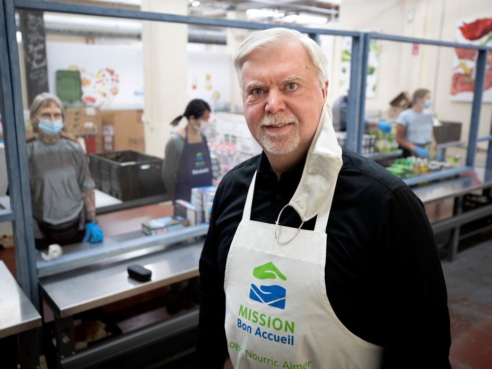 A man wears an apron reading Mission Bon Accueuil in a shelter's kitchen. Clients are seen waiting behind partitions in the background.