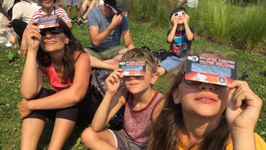 A mother and two children watch an eclipse through special glasses in a field.