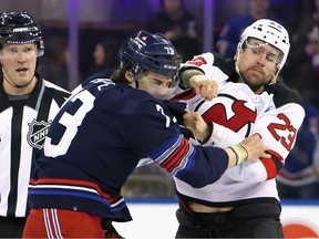 Devils' Kurtis MacDermid, right, is dressed in the Devils' white jersey and is seen throwing a right cross past the head of the Rangers' Matt Rempe whil ehis left hand is grabbing Rempe'sjersey. Both players are wearing helmets with visors on them. A linesman can be seen in the background watching the action unfold.