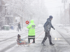 A crossing guard stops traffic as a person pulls a child in a sled across a street in the snow.