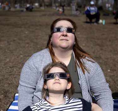 A woman and child wear eclipse glasses while sitting on the ground and looking at the sky.