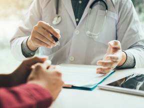 A doctor at a desk, seen from the neck down, with a stethoscope around their neck. A patient's hands are at bottom left.