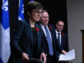 Three people in dark suits with a Quebec flag behind them are about to sit at a press conference.