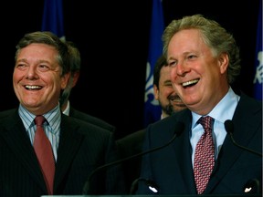Two men in suits and ties smile in front of a Quebec flag background during a press conference