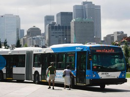 Two people stand next to an articulated bus. The Montreal skyline is in the background.