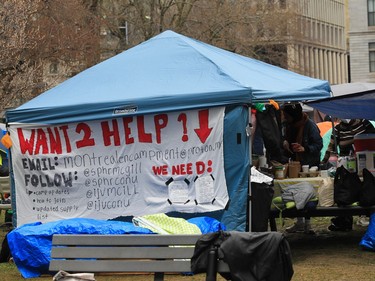 A sign with contact information asking for volunteers is visible on the side of a tent serving food in an encampment
