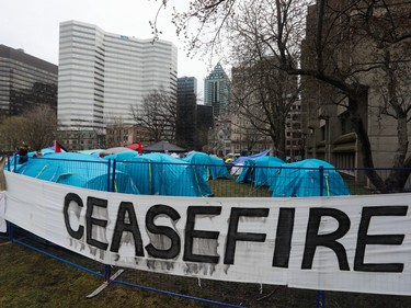 A sign reading 'CEASEFIRE!' is affixed to a temporary fence with tents behind it on a grassy area downtown