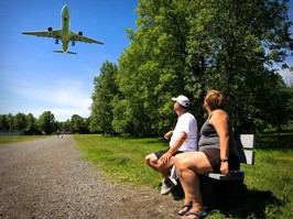 Two people sit on a bench and look at a plane flying overhead.