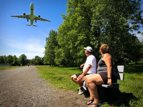 Two people sit on a bench and look at a plane flying overhead.