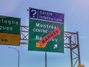 A highway sign showing the exit for Guy St. has an orange sticker saying barrée on top of it