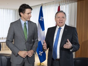 Prime Minister Justin Trudeau stands next to Quebec Premier François Legault who is talking and gesturing with his hands. There are Quebec and Canadian flags in the background.